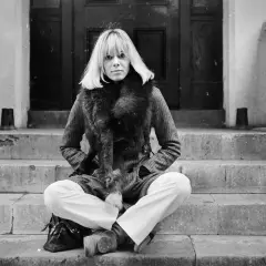 Anita Pallenberg, icona di stile, posa con un cappotto di tweed e una pelliccia voluminosa, seduta sulle scale di un portone.