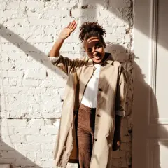 Donna con trench beige e pantaloni marroni, con la mano alzata per proteggersi dal sole. Un outfit versatile, ma quali scarpe abbinare?