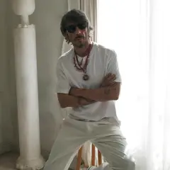 Pierpaolo Piccioli, con occhiali da sole e collane, posa con le braccia conserte in un ambiente chiaro con tende e colonne.