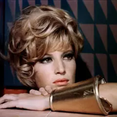 Monica Vitti, con un bracciale dorato e un'espressione pensierosa, sfoggia un look audace con capelli biondi cotonati e trucco marcato.
