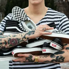 Un ragazzo con tatuaggi colorati tiene in braccio diversi tipi di Vans: Sk8-Hi, Slip-On a scacchi e altre classiche.