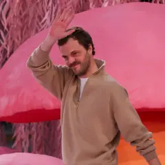 Matthieu Blazy saluta con la mano, sorridendo, su uno sfondo rosa con elementi che ricordano funghi e piante.