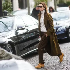 Donna con un elegante outfit con Timberland, cappotto verde oliva, dolcevita bianco e pantaloni marroni, cammina su un acciottolato.