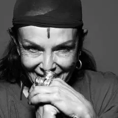 Michele Lamy, con un turbante nero e un segno sulla fronte, sorride enigmaticamente, le mani adornate da anelli vistosi.