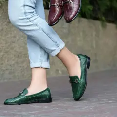 Un paio di jeans chiari e un paio di eleganti mocassini uomo verdi con fibbia metallica. Un altro paio di mocassini marroni è tenuto in mano.