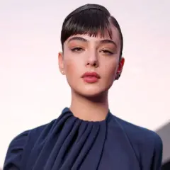 Primo piano di Deva Cassel con un'espressione intensa, indossa un abito blu scuro con un collo drappeggiato.