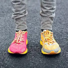 Due diversi tipi di scarpe da ginnastica, una rosa acceso e una arancione, indossate con pantaloni grigi su una superficie scura.