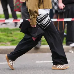 Persona con un outfit casual, pantaloni neri ampi, giacca color kaki e scarpe da ginnastica Adidas marroni.