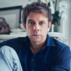 Ritratto di Jonathan Anderson, con occhi azzurri penetranti, indossa una camicia blu e jeans.