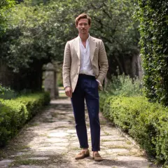 Uomo con giacca beige e pantaloni blu, un look che incarna lo stile francese uomo, in un giardino lussureggiante.
