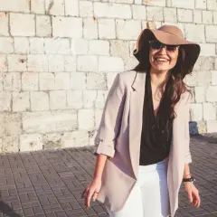 Donna sorridente con cappello e occhiali da sole, perfetta per come vestirsi con 20 gradi: blazer leggero, top nero e pantaloni bianchi.