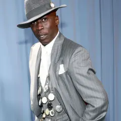 Un uomo con un cappello grigio e un completo gessato, ornato da numerosi orologi da taschino, incarna l'eleganza maschile.
