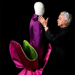 Roberto Capucci, maestro della moda, accarezza un abito viola su un manichino, circondato da petali fucsia e verdi.