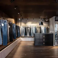 Jeans appesi in un negozio, con una **gamba affusolata** che si nota in primo piano. L'ambiente è moderno, con pavimenti in legno scuro e illuminazione a faretti.