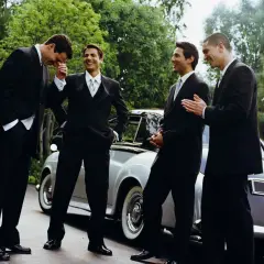 Quattro uomini in abito scuro, perfetti per un matrimonio, ridono vicino a un'auto d'epoca.