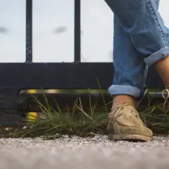 Gambe con jeans arrotolati e scarpe senza calzini, appoggiate su un muretto con erba e ghiaia.