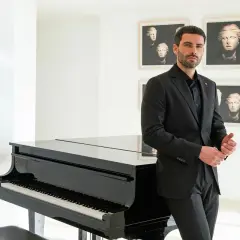 Uomo in elegante outfit teatro, con completo nero, accanto a un pianoforte a coda.