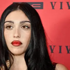 Lourdes Leon, con labbra rosse audaci e sguardo intenso, posa davanti a uno sfondo rosso con scritte nere.
