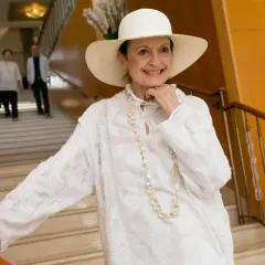 Carla Fracci sorride con un cappello bianco e una collana di perle, elegante su una scala.