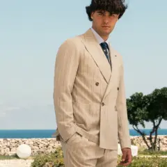 Uomo elegante con abito gessato beige, in piedi con lo sguardo rivolto verso l'osservatore, con il mare e il cielo azzurro sullo sfondo.