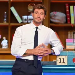 Stefano De Martino outfit: camicia bianca, cravatta blu scuro e pantaloni eleganti. Sullo sfondo, una libreria piena di oggetti colorati.