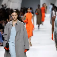 Angelina Kendall sfila con un cappotto grigio oversize e un abito a righe azzurre, guanti arancioni vivaci.
