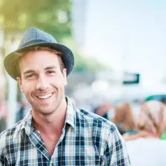 Uomo sorridente con un cappello adatto al viso, indossa una camicia a quadri. La luce del sole illumina la scena.