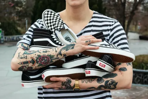 Un ragazzo con tatuaggi colorati tiene in braccio diversi tipi di Vans: Sk8-Hi, Slip-On a scacchi e altre classiche.