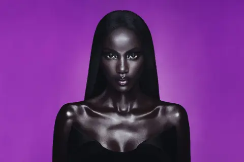 Anok Yai, con pelle lucida e capelli neri, posa con uno sguardo intenso su sfondo viola.