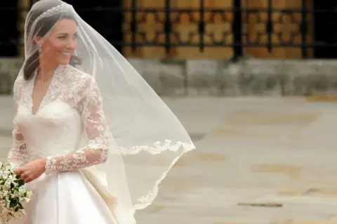 Kate Middleton sposa, radiosa nel suo abito bianco con pizzo e velo, tiene un bouquet di fiori.