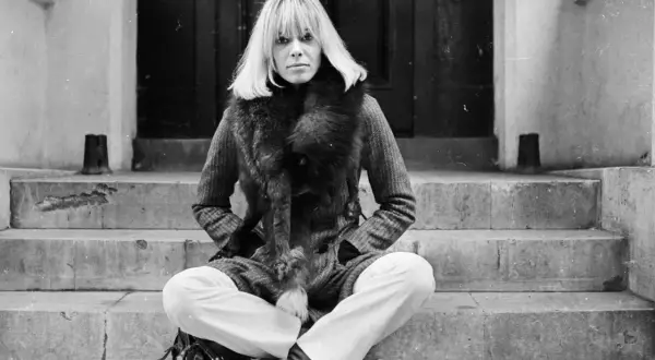 Anita Pallenberg, icona di stile, posa con un cappotto di tweed e una pelliccia voluminosa, seduta sulle scale di un portone.