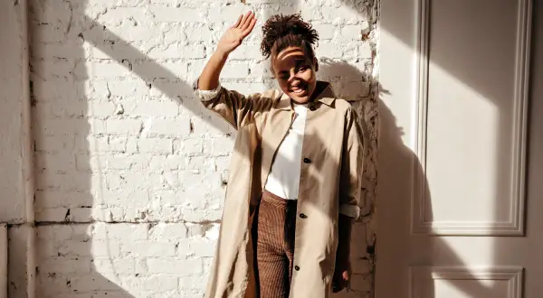 Donna con trench beige e pantaloni marroni, con la mano alzata per proteggersi dal sole. Un outfit versatile, ma quali scarpe abbinare?