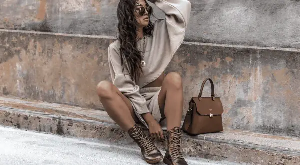 Donna con un maglione corto e pantaloncini beige, stivali stringati e borsa marrone. Sembra un uomo basso.