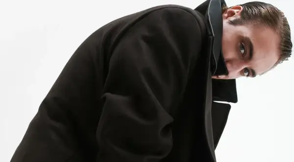 Luca Vergoni, con un cappotto scuro e un'espressione enigmatica, guarda verso l'obiettivo.