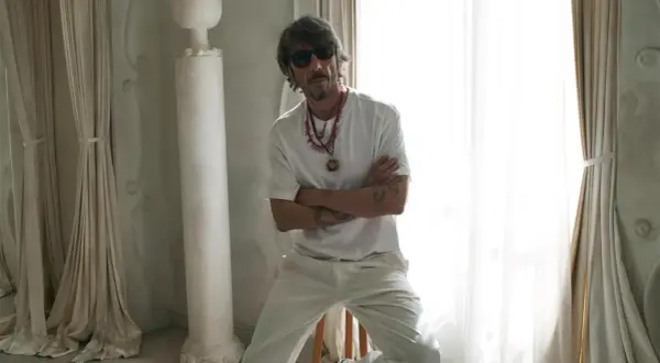 Pierpaolo Piccioli, con occhiali da sole e collane, posa con le braccia conserte in un ambiente chiaro con tende e colonne.