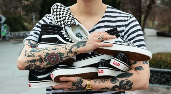 Un ragazzo con tatuaggi colorati tiene in braccio diversi tipi di Vans: Sk8-Hi, Slip-On a scacchi e altre classiche.