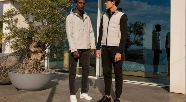 Due uomini con un outfit uomo elegante sportivo, uno con giacca grigia e jeans, l'altro con gilet grigio e pantaloni neri, in piedi all'esterno.