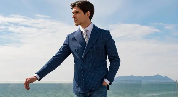 Uomo con giacca blu a doppio petto a righe, camicia bianca e cravatta, appoggiato a una ringhiera con vista mare.