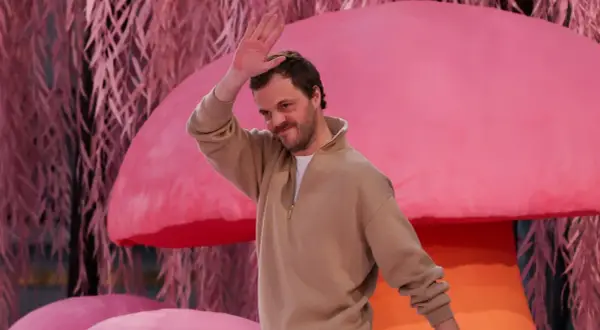 Matthieu Blazy saluta con la mano, sorridendo, su uno sfondo rosa con elementi che ricordano funghi e piante.