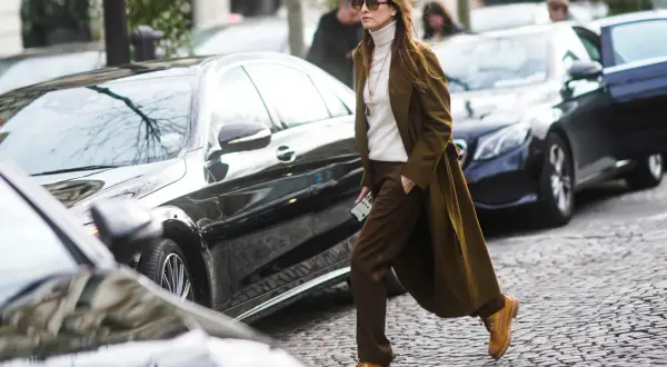 Donna con un elegante outfit con Timberland, cappotto verde oliva, dolcevita bianco e pantaloni marroni, cammina su un acciottolato.