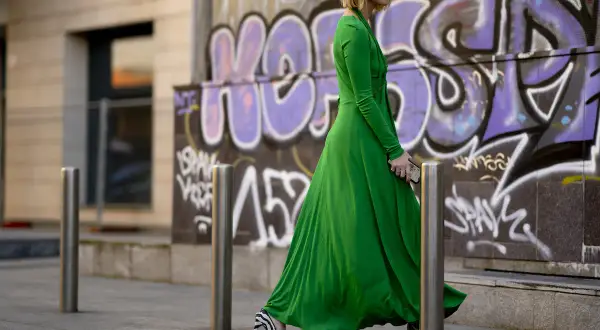 Donna con vestito lungo elegante verde smeraldo e scarpe zebrate, cammina con passo deciso davanti a un muro con graffiti.