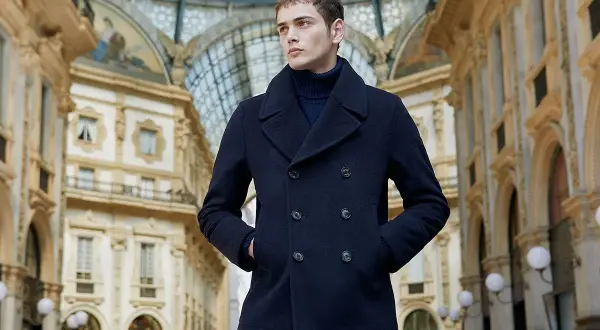 Giovane uomo con un caban blu scuro, un capo che incarna eleganza e calore, passeggia sotto la volta di una galleria storica.