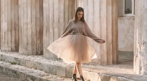 Ragazza con un outfit gonna tulle lunga color cipria, maglione oversize e anfibi, passeggia tra colonne antiche.
