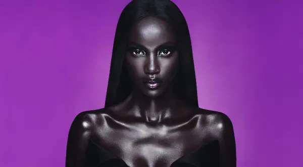 Anok Yai, con pelle lucida e capelli neri, posa con uno sguardo intenso su sfondo viola.