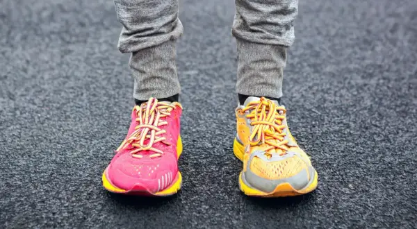 Due diversi tipi di scarpe da ginnastica, una rosa acceso e una arancione, indossate con pantaloni grigi su una superficie scura.