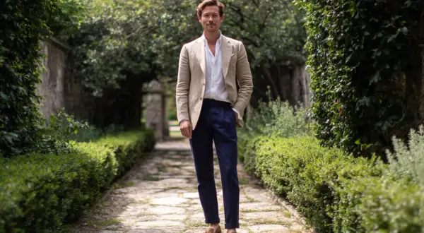 Uomo con giacca beige e pantaloni blu, un look che incarna lo stile francese uomo, in un giardino lussureggiante.