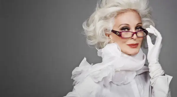 Carmen Dell'Orefice, icona di stile, con capelli bianchi voluminosi, occhiali viola e guanti bianchi, posa con eleganza.