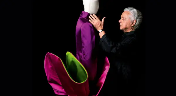 Roberto Capucci, maestro della moda, accarezza un abito viola su un manichino, circondato da petali fucsia e verdi.