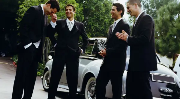 Quattro uomini in abito scuro, perfetti per un matrimonio, ridono vicino a un'auto d'epoca.