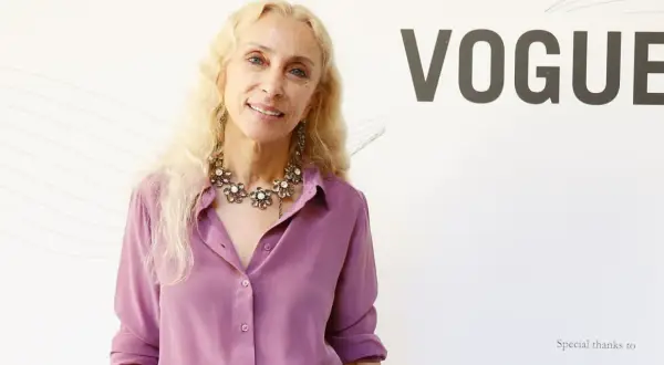Franca Sozzani, icona di stile, sorride con un collier floreale e una camicia lilla, sullo sfondo del logo VOGUE.